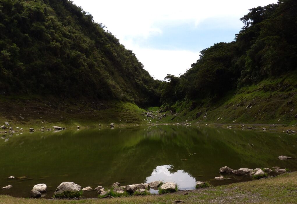 Santuario Nacional de Ampay: laguna de agua verde y quieta rodeada de piedras, al pie de dos laderas cubiertas de bosque denso bajo un cielo nublado