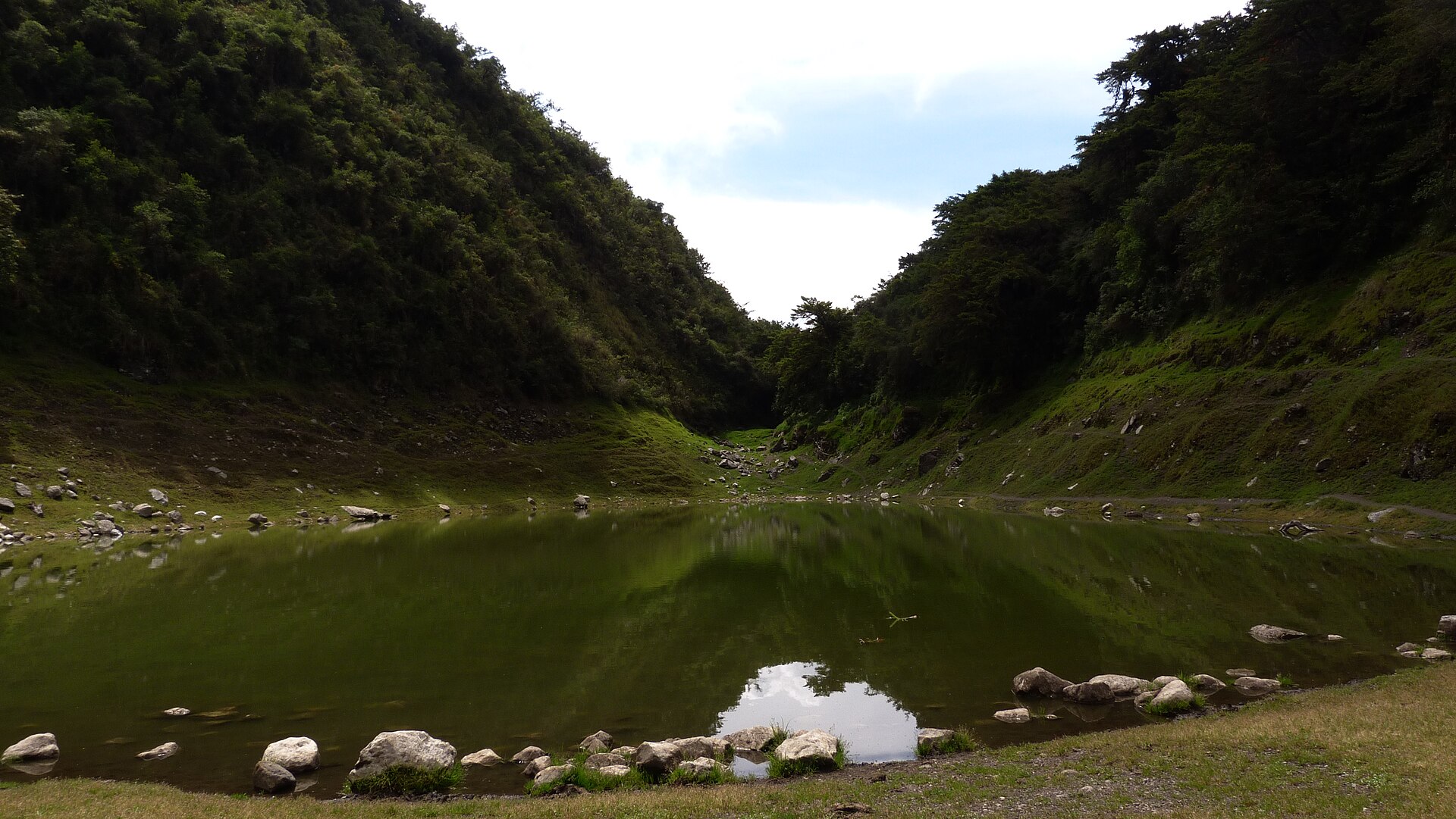 Santuario Nacional de Ampay: laguna de agua verde y quieta rodeada de piedras, al pie de dos laderas cubiertas de bosque denso bajo un cielo nublado