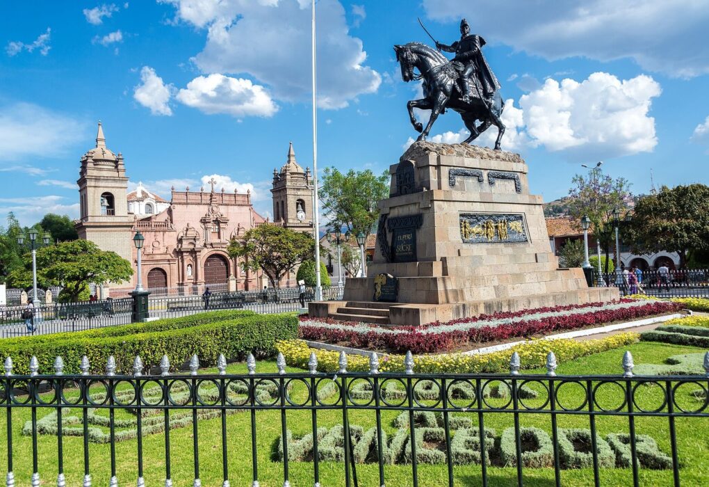Plaza de Armas de Ayacucho de día, con el monumento a Sucre, la Catedral al fondo a la izquierda y un jardín con flores bajo un cielo azul con nubes