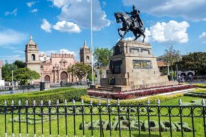 Plaza de Armas de Ayacucho de día, con el monumento a Sucre, la Catedral al fondo a la izquierda y un jardín con flores bajo un cielo azul con nubes