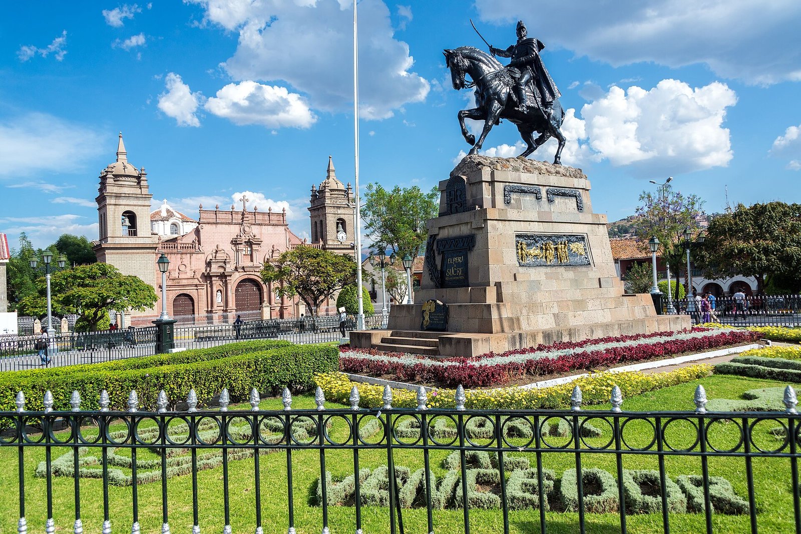 Plaza de Armas de Ayacucho de día, con el monumento a Sucre, la Catedral al fondo a la izquierda y un jardín con flores bajo un cielo azul con nubes