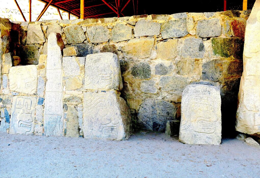 Casma: monolitos de Cerro Sechín con rostros y figuras talladas, frente a un muro de piedras bajo un techo protector