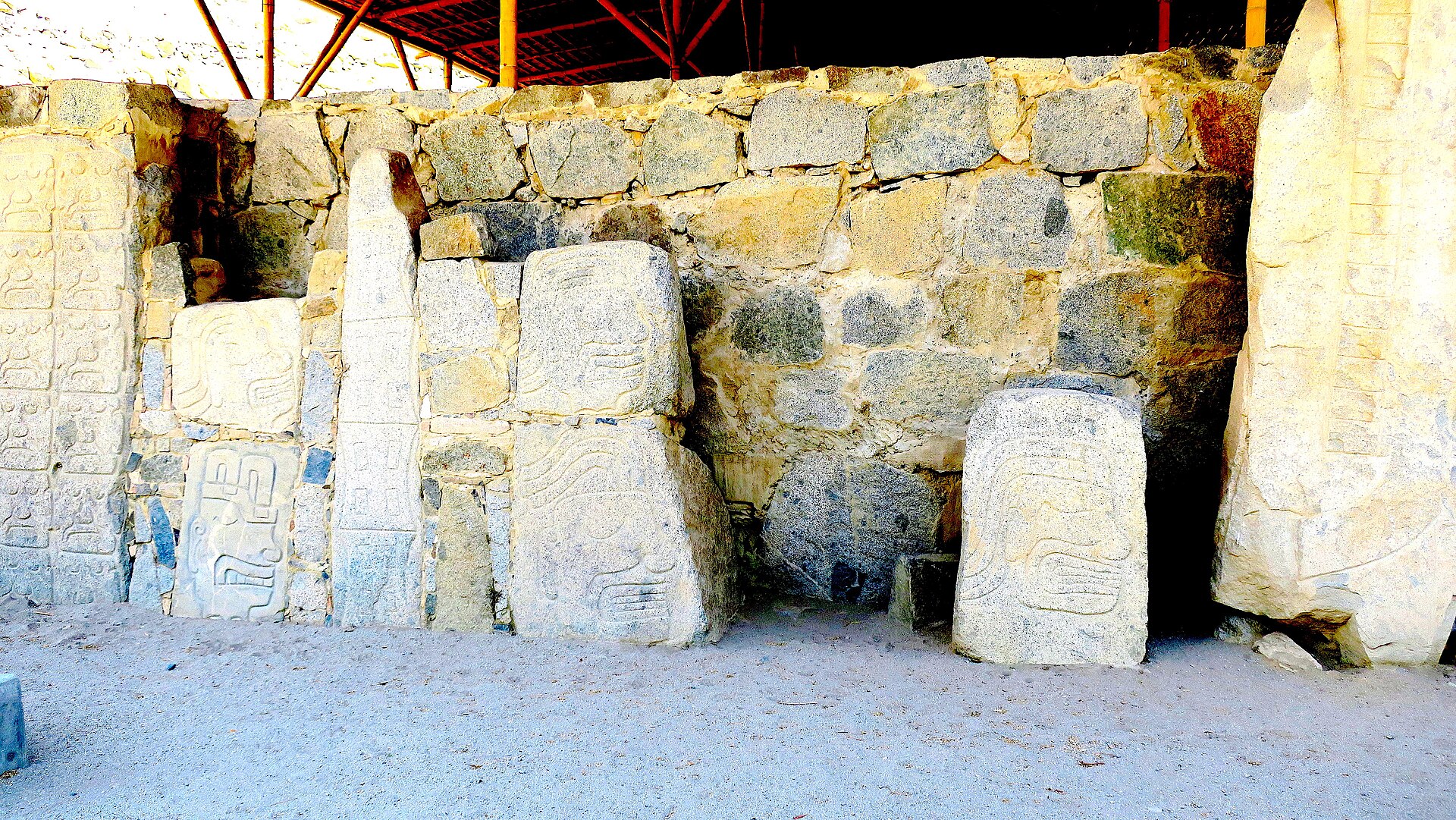 Casma: monolitos de Cerro Sechín con rostros y figuras talladas, frente a un muro de piedras bajo un techo protector
