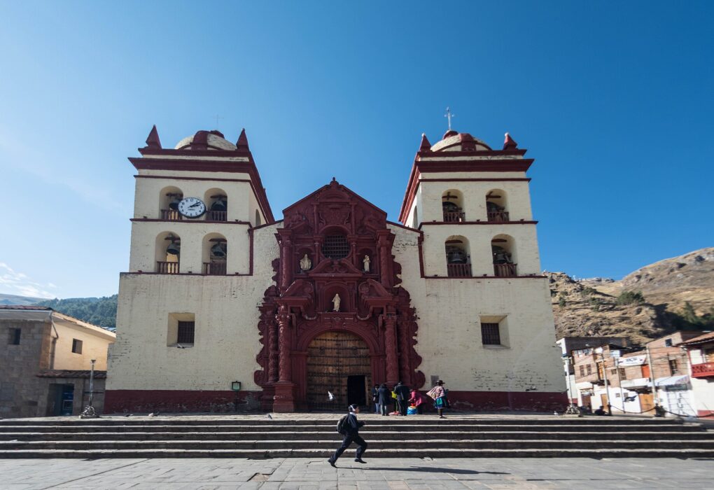 Catedral de Huancavelica, de fachada blanca con portada barroca rojiza y dos torres con campanas, frente a una escalinata, bajo un cielo azul