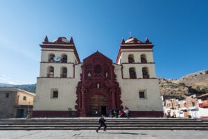 Catedral de Huancavelica, de fachada blanca con portada barroca rojiza y dos torres con campanas, frente a una escalinata, bajo un cielo azul