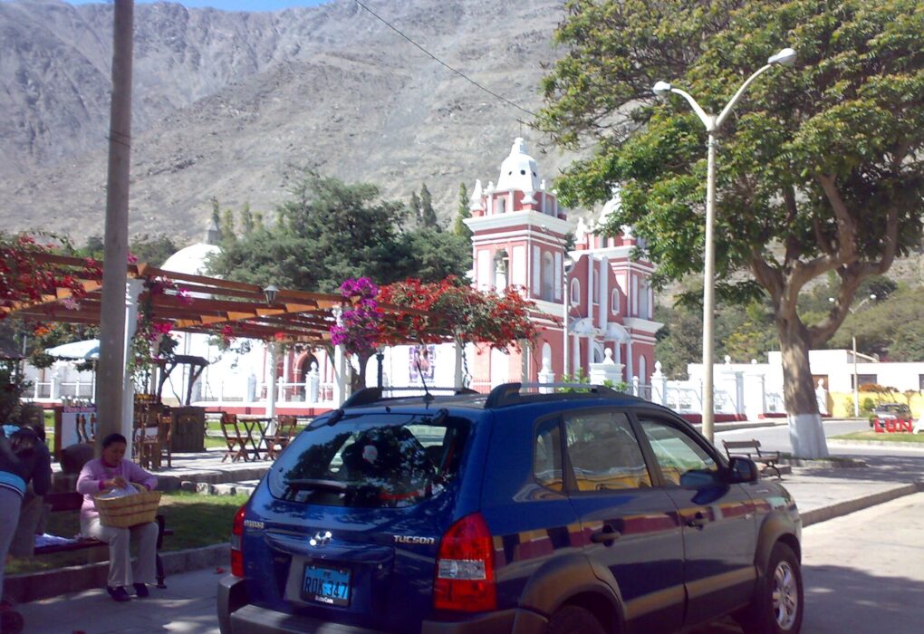 Plaza de Lunahuaná con iglesia de fachada roja y dos torres blancas, buganvilias y un cerro seco al fondo