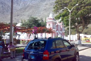 Plaza de Lunahuaná con iglesia de fachada roja y dos torres blancas, buganvilias y un cerro seco al fondo