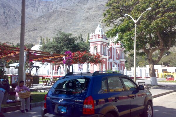 Plaza de Lunahuaná con iglesia de fachada roja y dos torres blancas, buganvilias y un cerro seco al fondo
