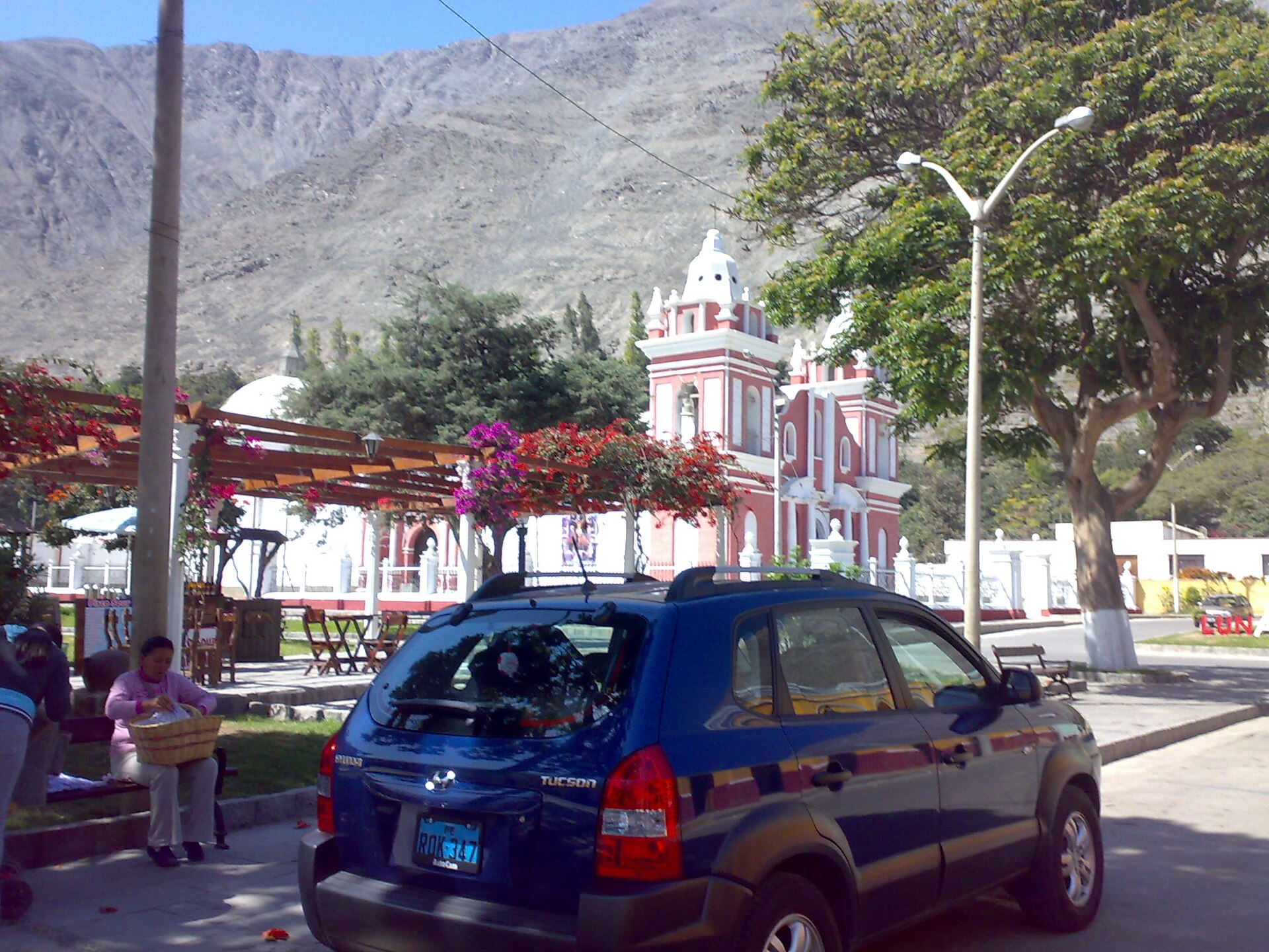 Plaza de Lunahuaná con iglesia de fachada roja y dos torres blancas, buganvilias y un cerro seco al fondo