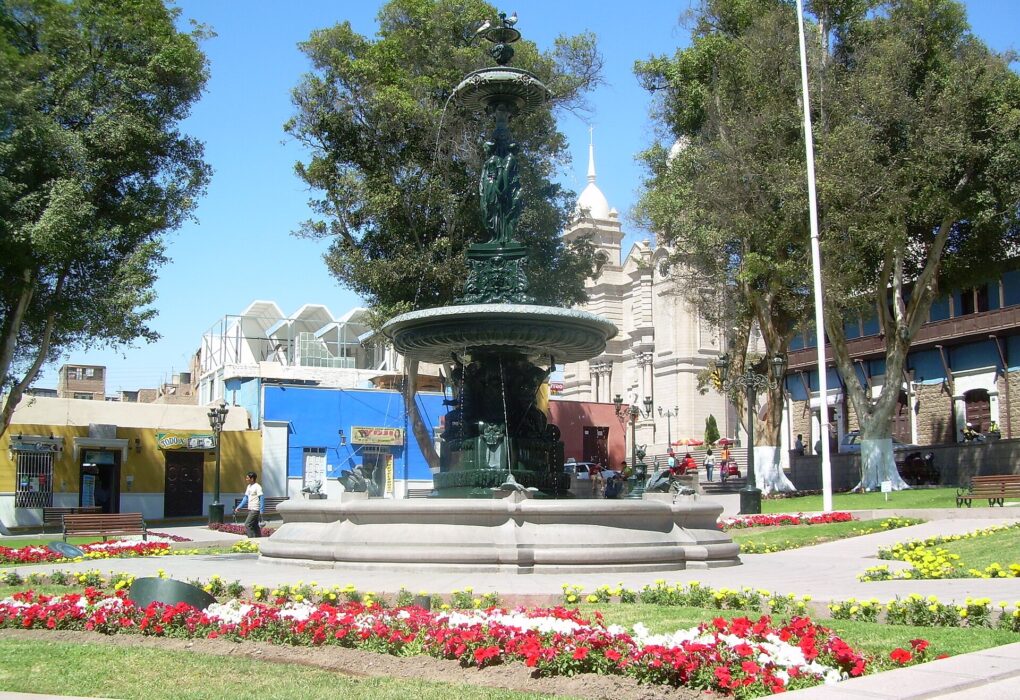 Plaza Mayor de Moquegua con su fuente de bronce, jardines con flores rojas y blancas, árboles y la torre blanca de una iglesia al fondo