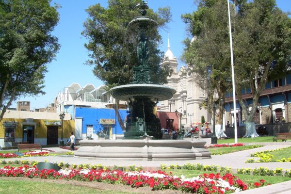 Plaza Mayor de Moquegua con su fuente de bronce, jardines con flores rojas y blancas, árboles y la torre blanca de una iglesia al fondo