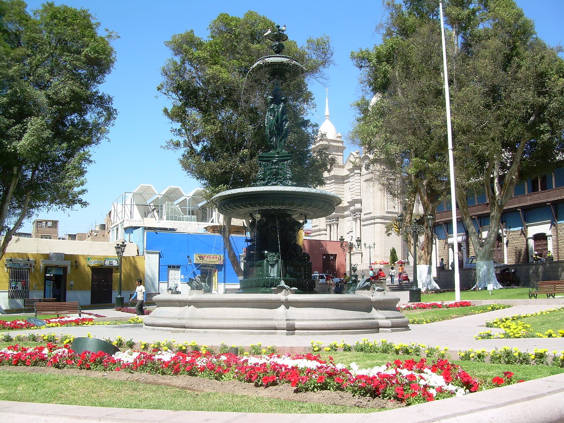 Plaza Mayor de Moquegua con su fuente de bronce, jardines con flores rojas y blancas, árboles y la torre blanca de una iglesia al fondo