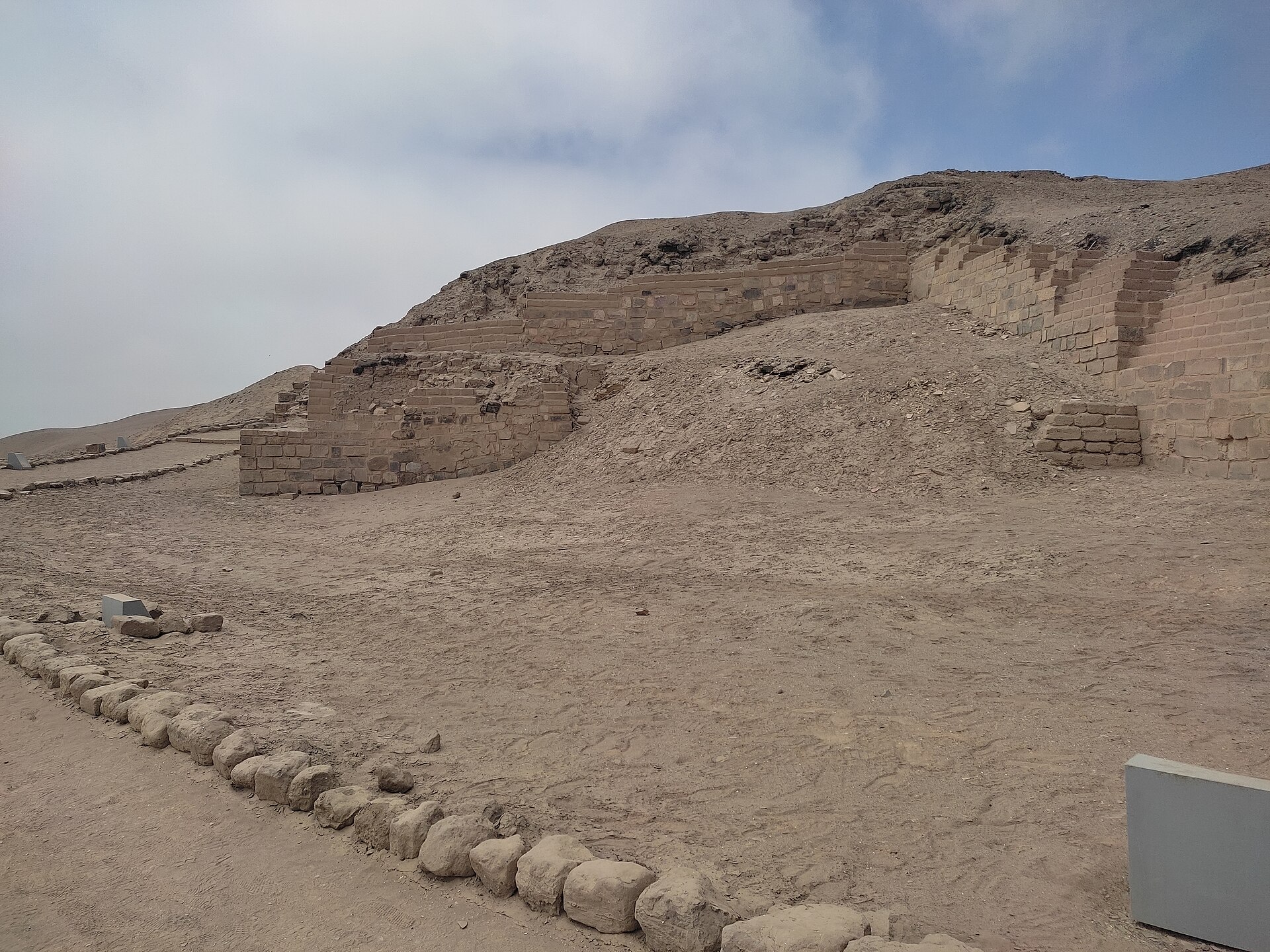 Pachacamac: restos del Templo del Sol, muros de adobe escalonados sobre una loma de tierra, con explanada y piedras alineadas bajo cielo nublado