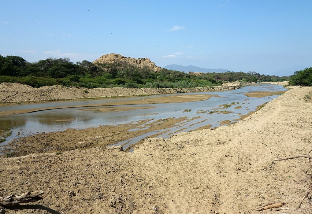Bosque de Pómac: río de poco caudal entre orillas de arena, con árboles verdes y una gran huaca de adobe erosionada al fondo bajo un cielo azul
