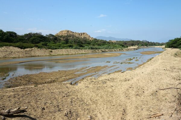 Bosque de Pómac: río de poco caudal entre orillas de arena, con árboles verdes y una gran huaca de adobe erosionada al fondo bajo un cielo azul