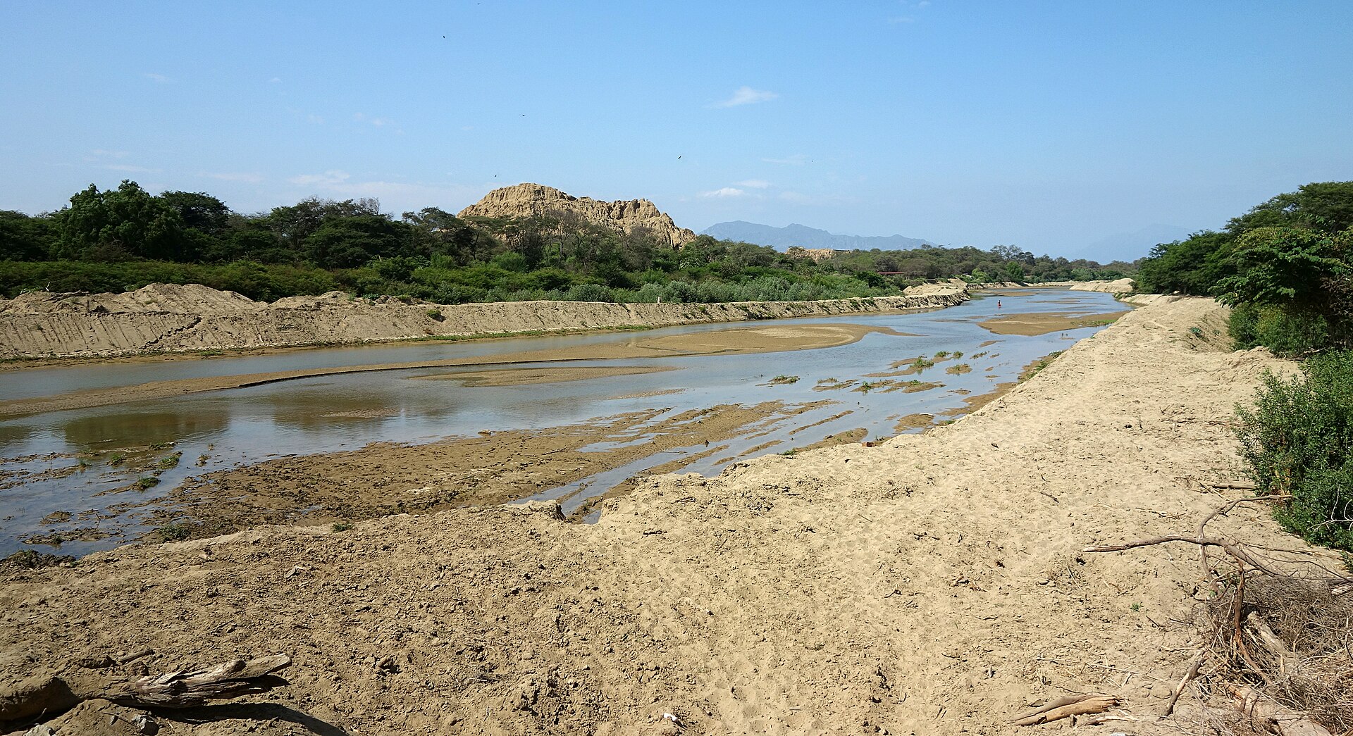 Bosque de Pómac: río de poco caudal entre orillas de arena, con árboles verdes y una gran huaca de adobe erosionada al fondo bajo un cielo azul