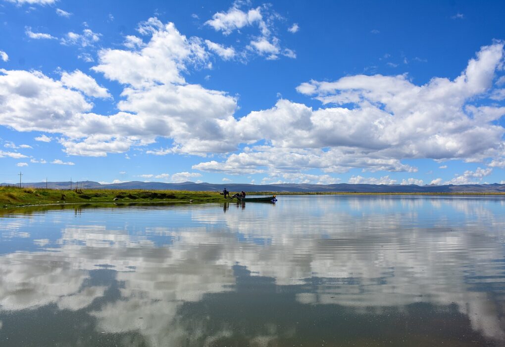 Reserva Nacional de Junín: el lago Chinchaycocha en calma refleja nubes blancas y cielo azul, con una orilla baja de pastos y un bote pequeño a lo lejos