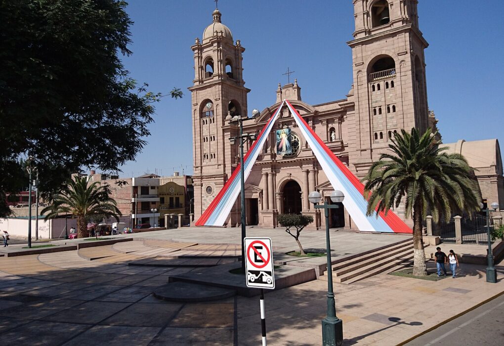 Catedral de Tacna, de piedra rosada y con dos torres, con cintas rojas y blancas en la fachada y una plaza con palmeras al frente