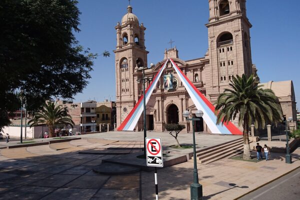 Catedral de Tacna, de piedra rosada y con dos torres, con cintas rojas y blancas en la fachada y una plaza con palmeras al frente