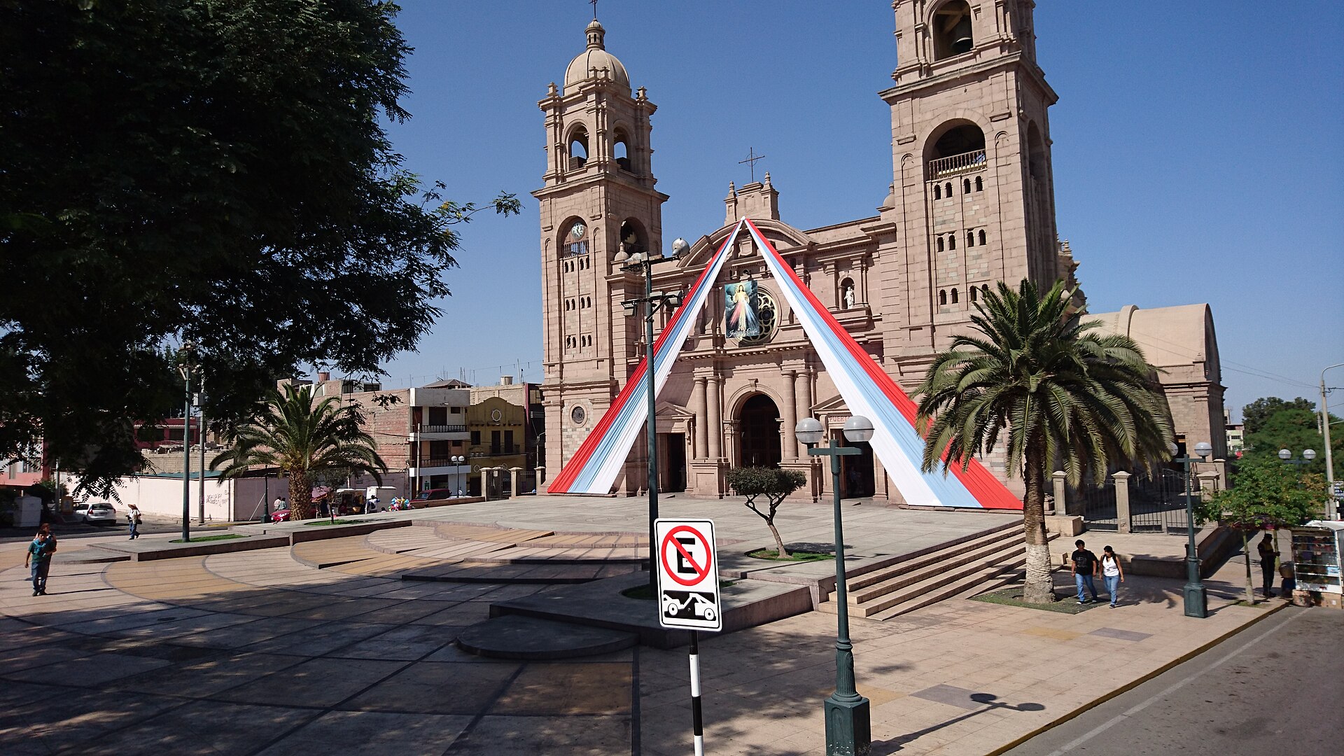 Catedral de Tacna, de piedra rosada y con dos torres, con cintas rojas y blancas en la fachada y una plaza con palmeras al frente